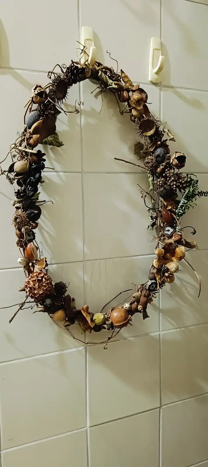 A dried autumn wreath made of various seeds, pods, and twigs hangs on a white tiled wall using two white hooks. The wreath…