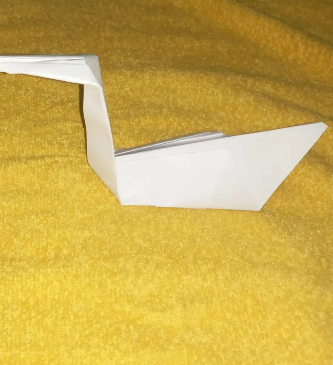 A white origami swan rests on a textured yellow fabric surface. The swan is made from folded paper with a long neck and a…