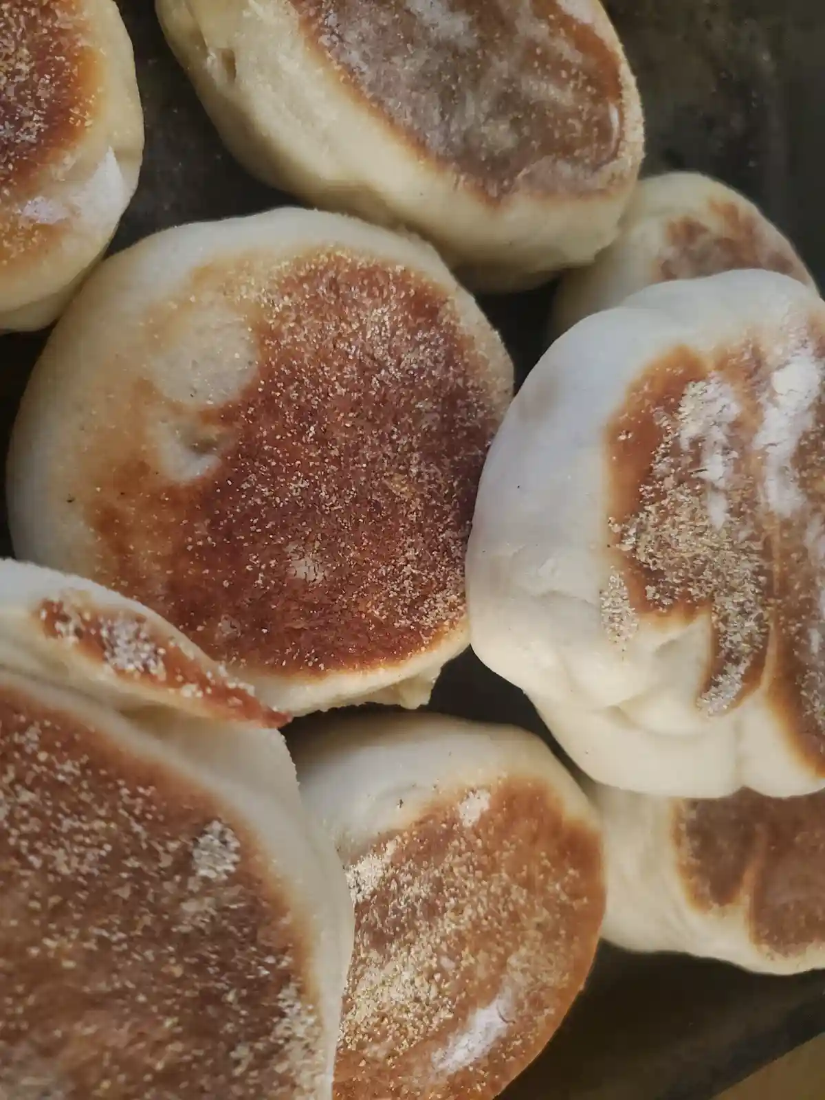 A close-up photo shows several golden-brown, toasted english muffins with visible flour dusting, arranged on a dark surface…