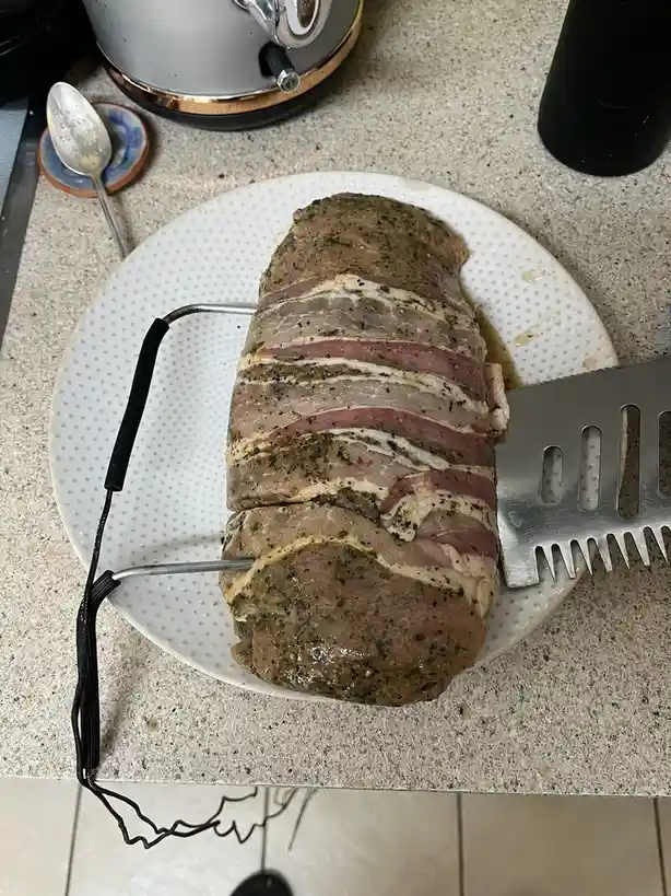 A raw, herb-seasoned beef roast wrapped in bacon sits on a white dotted plate, with a metal meat thermometer inserted and a…