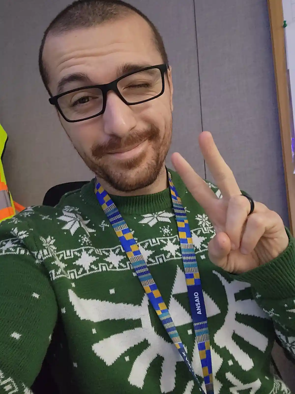 A man with glasses and a beard wears a green Christmas sweater with white snowflake patterns and makes a peace sign with his…