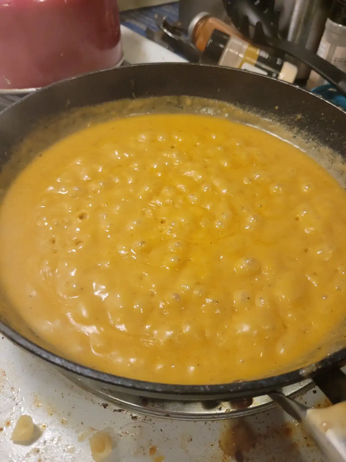 A close-up photo shows a bubbling, creamy orange macaroni and cheese dish simmering in a black frying pan on a stovetop. The…