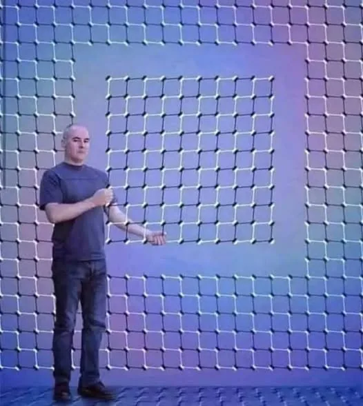 A bald man in a dark t-shirt and jeans stands in front of a large, glowing blue grid background. He gestures with his right…