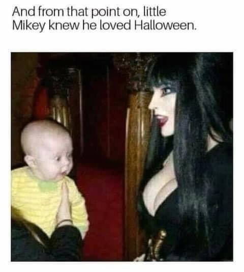 A baby in a yellow striped shirt looks up at a woman dressed as Elvira, Mistress of the Dark, who is leaning toward him with…