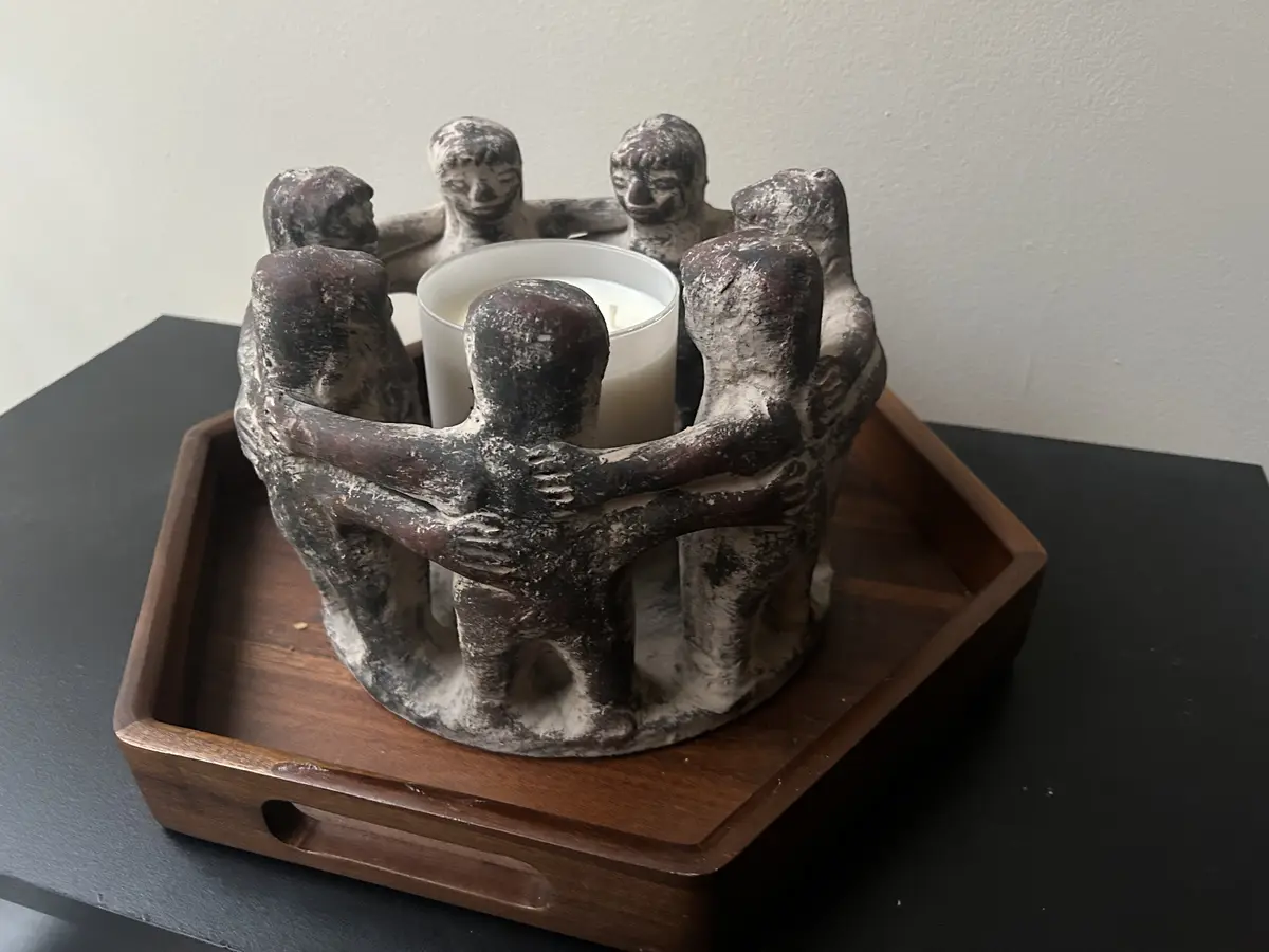 A white candle sits in a decorative holder shaped like six abstract human figures hugging in a circle, placed on a wooden…