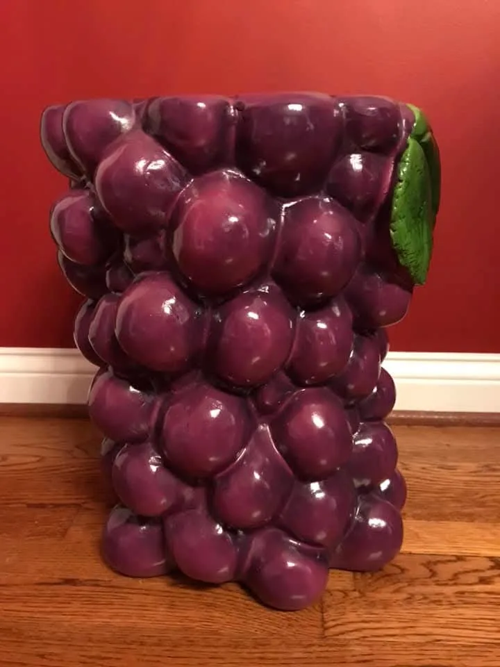 a glossy purple ceramic stool shaped like a bunch of grapes with a green leaf on the side, sitting on a wooden floor against…
