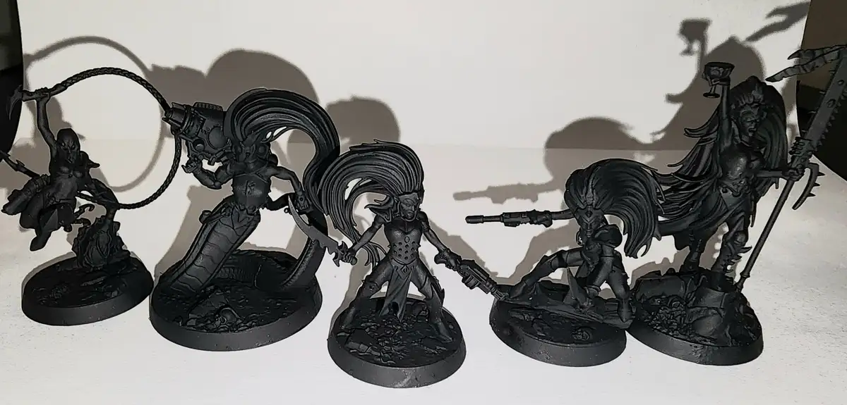 Five unpainted black miniature figurines of fantasy or sci-fi characters are lined up on a white surface, each holding…