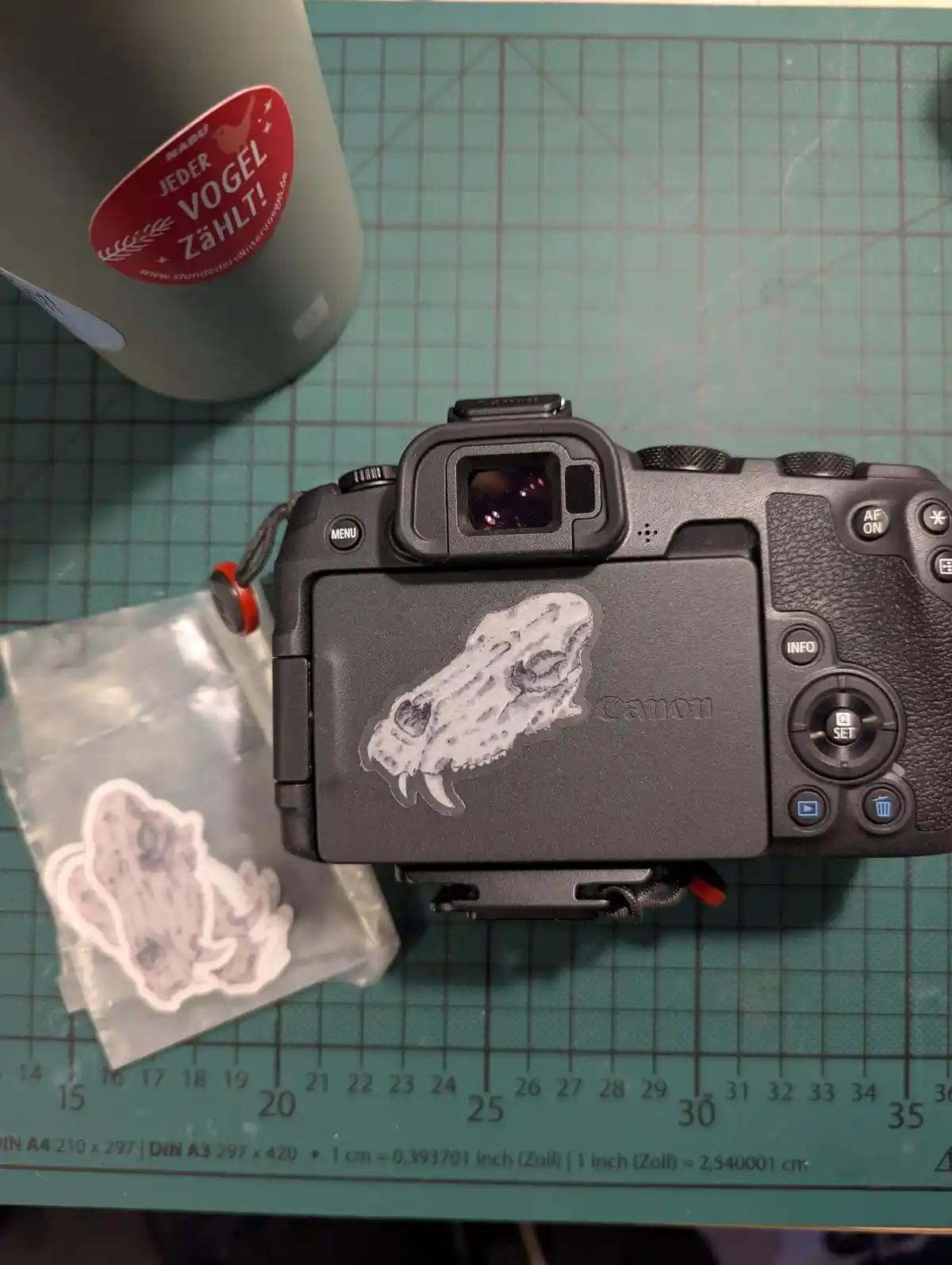 A black Canon camera sits on a green cutting mat, its back covered by a sticker of a stylized animal skull. Beside it is a…