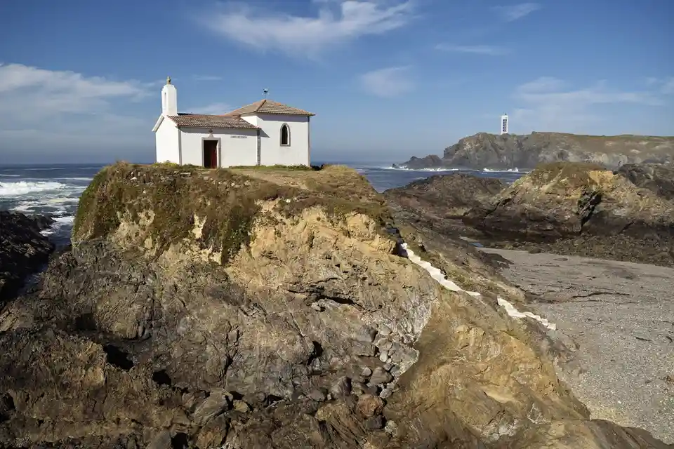 A scenic coastal photo shows a small white chapel perched on a rocky cliff overlooking the ocean, with a lighthouse visible…