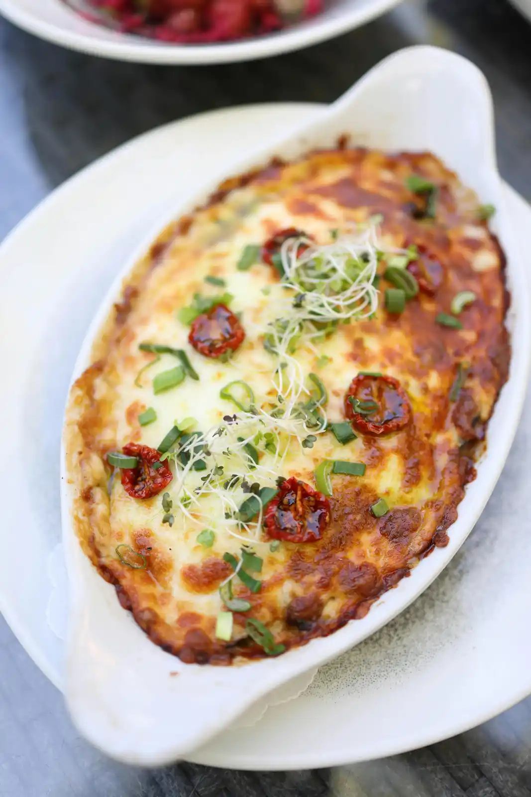 A close-up photo shows a white oval baking dish filled with a baked dish featuring a golden-brown, melted cheese topping…