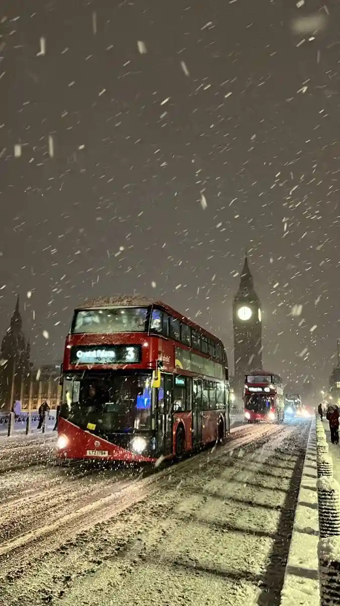 A red double-decker bus drives through heavy snowfall on a London street at night, with Big Ben visible in the background…