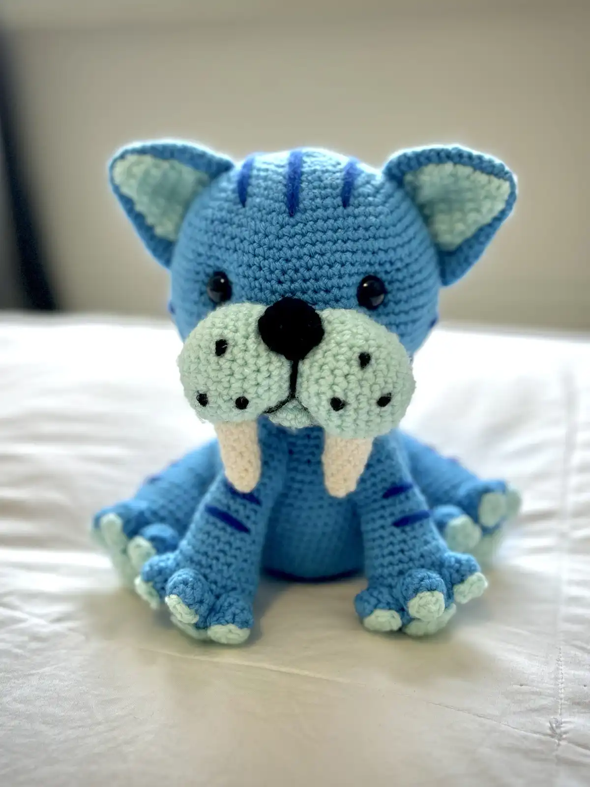 A handmade blue crocheted stuffed animal with cat-like ears, tiger stripes, and prominent walrus tusks, sitting on a white…