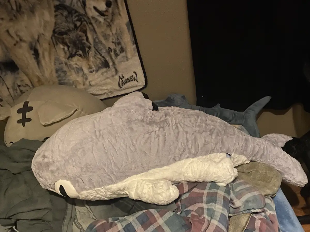 A large, light purple plush whale lies horizontally on a messy bed with blankets and other stuffed animals, including a…
