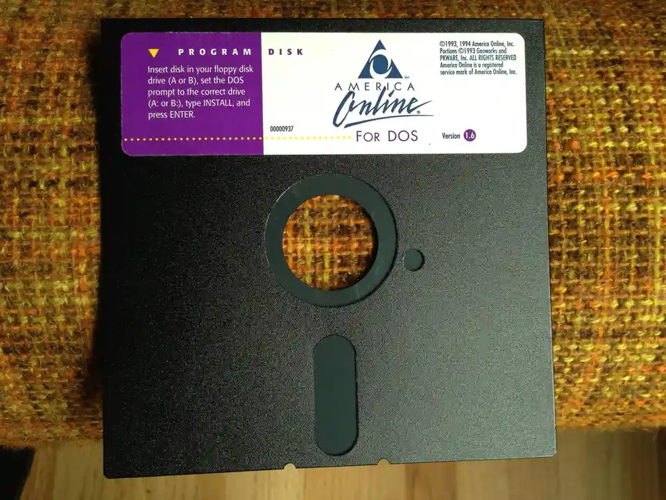 A black 3.5-inch floppy disk labeled 'America Online FOR DOS Version 1.6' sits on a textured orange and green fabric. The…
