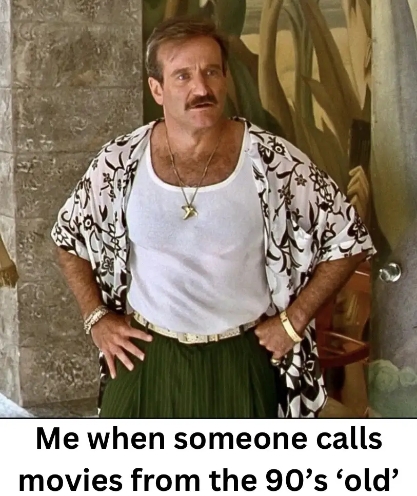 A man with a mustache, wearing a white tank top under a patterned open shirt and green pants, stands with hands on hips…