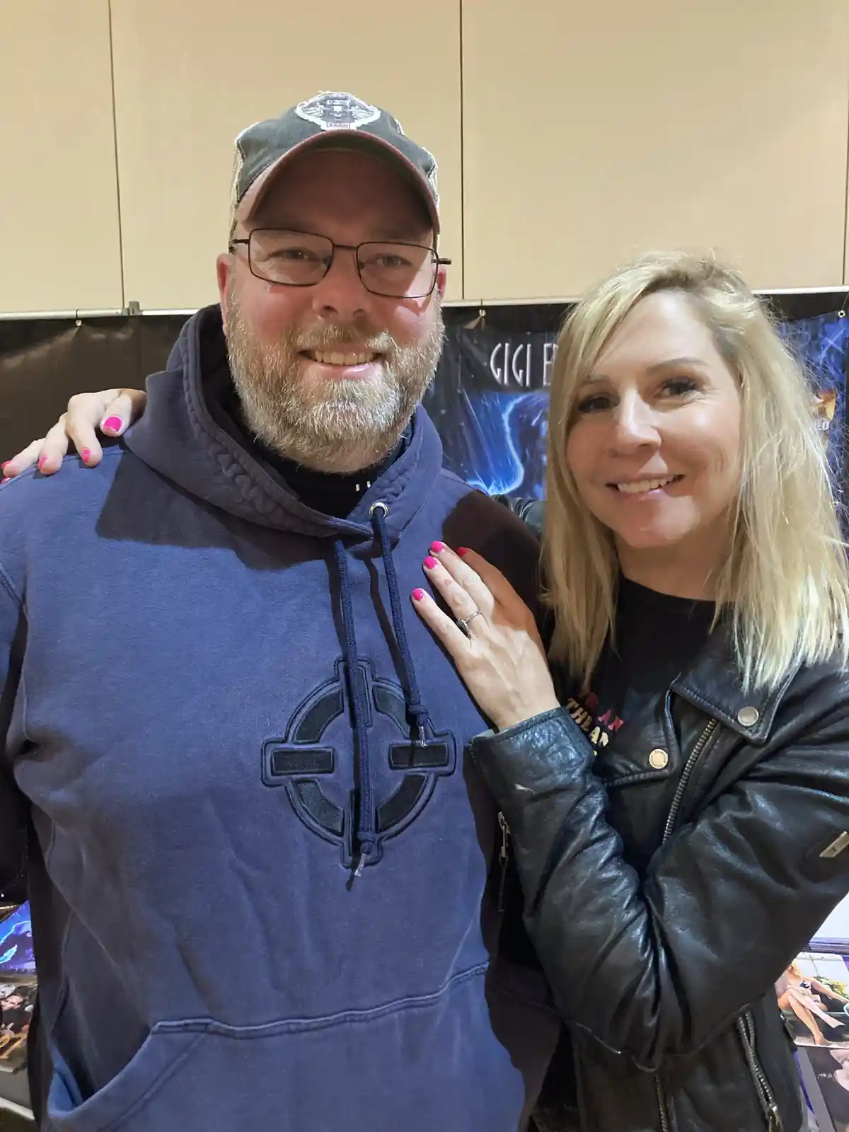 A man with glasses and a beard wearing a blue hoodie and cap stands smiling with a woman in a black leather jacket. Her…