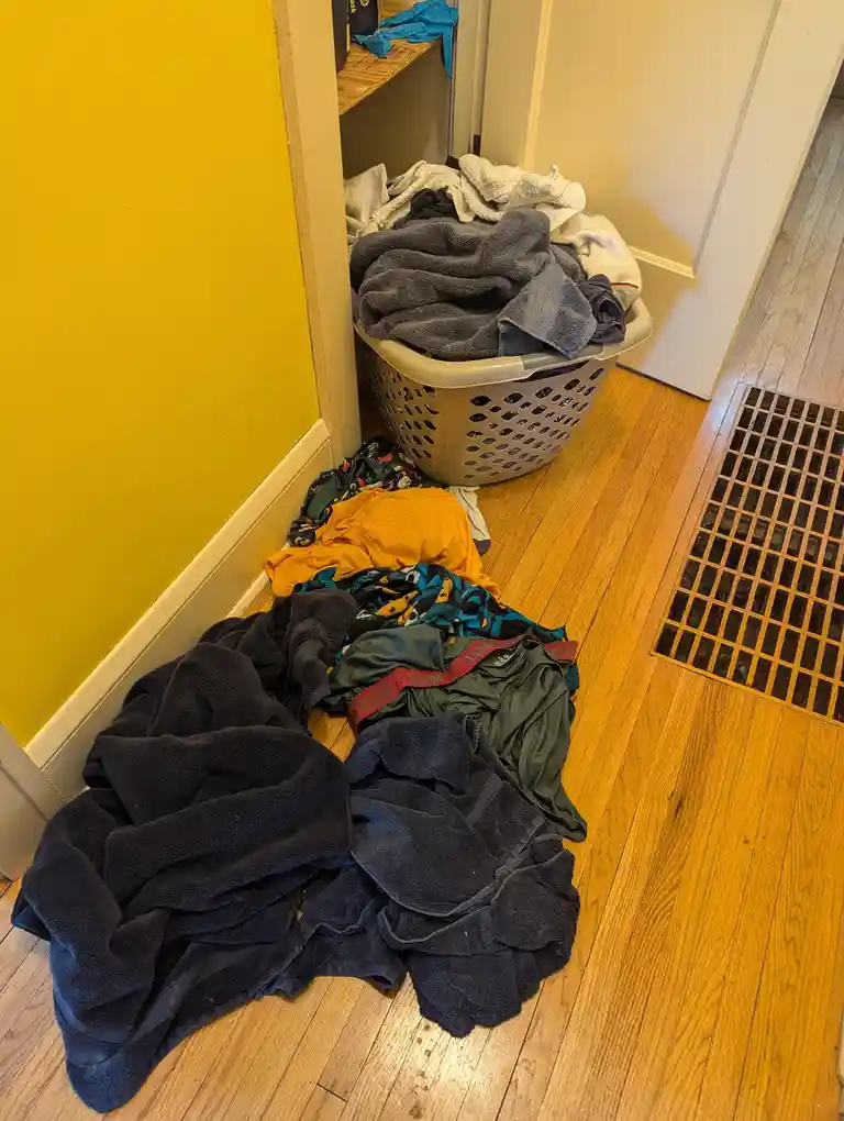 A messy laundry scene shows a beige plastic basket overflowing with clothes, placed in a corner next to a yellow wall and a…