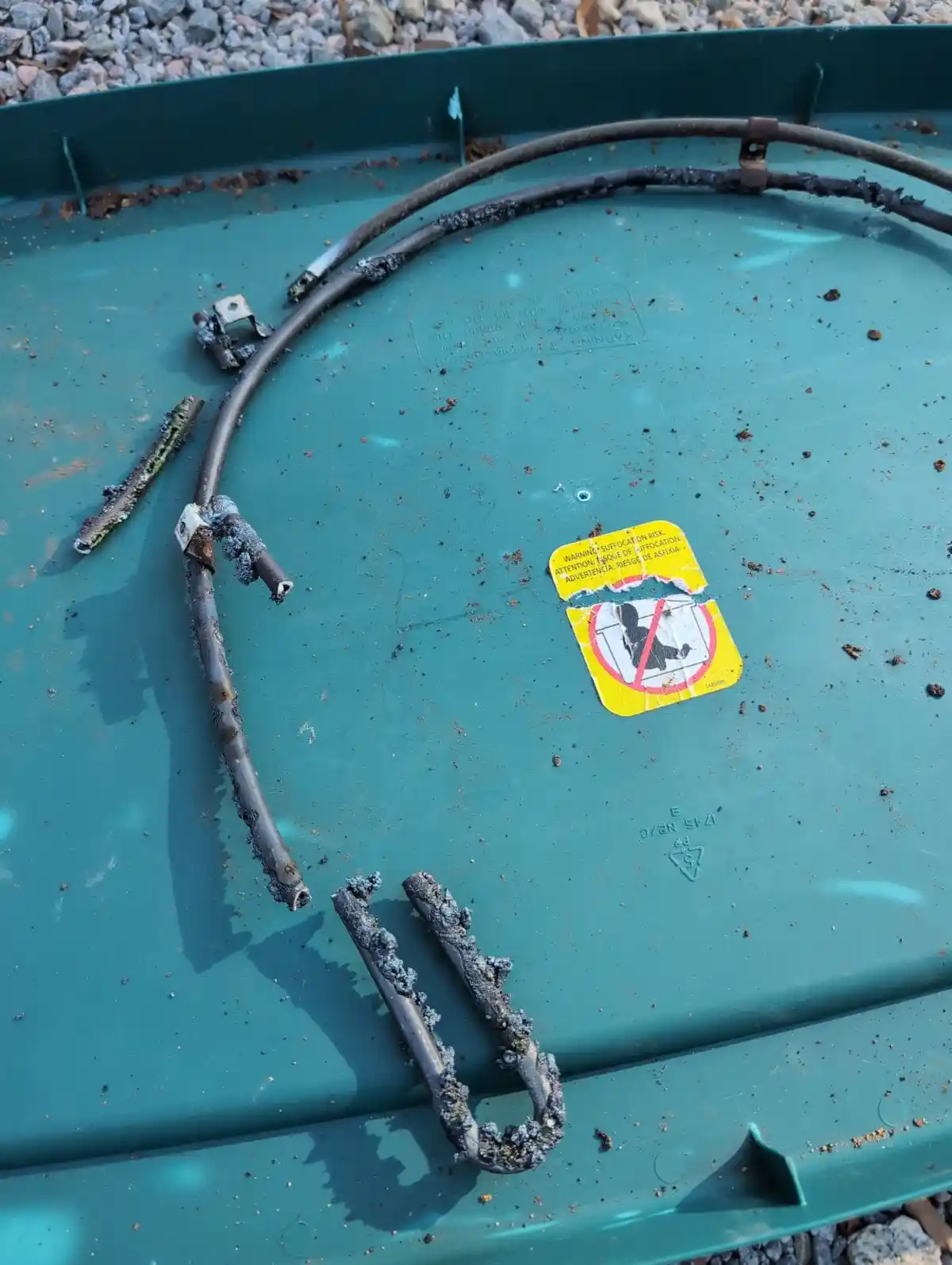 A close-up of a green plastic trash can lid covered in dirt and debris, featuring a torn yellow suffocation warning sticker…