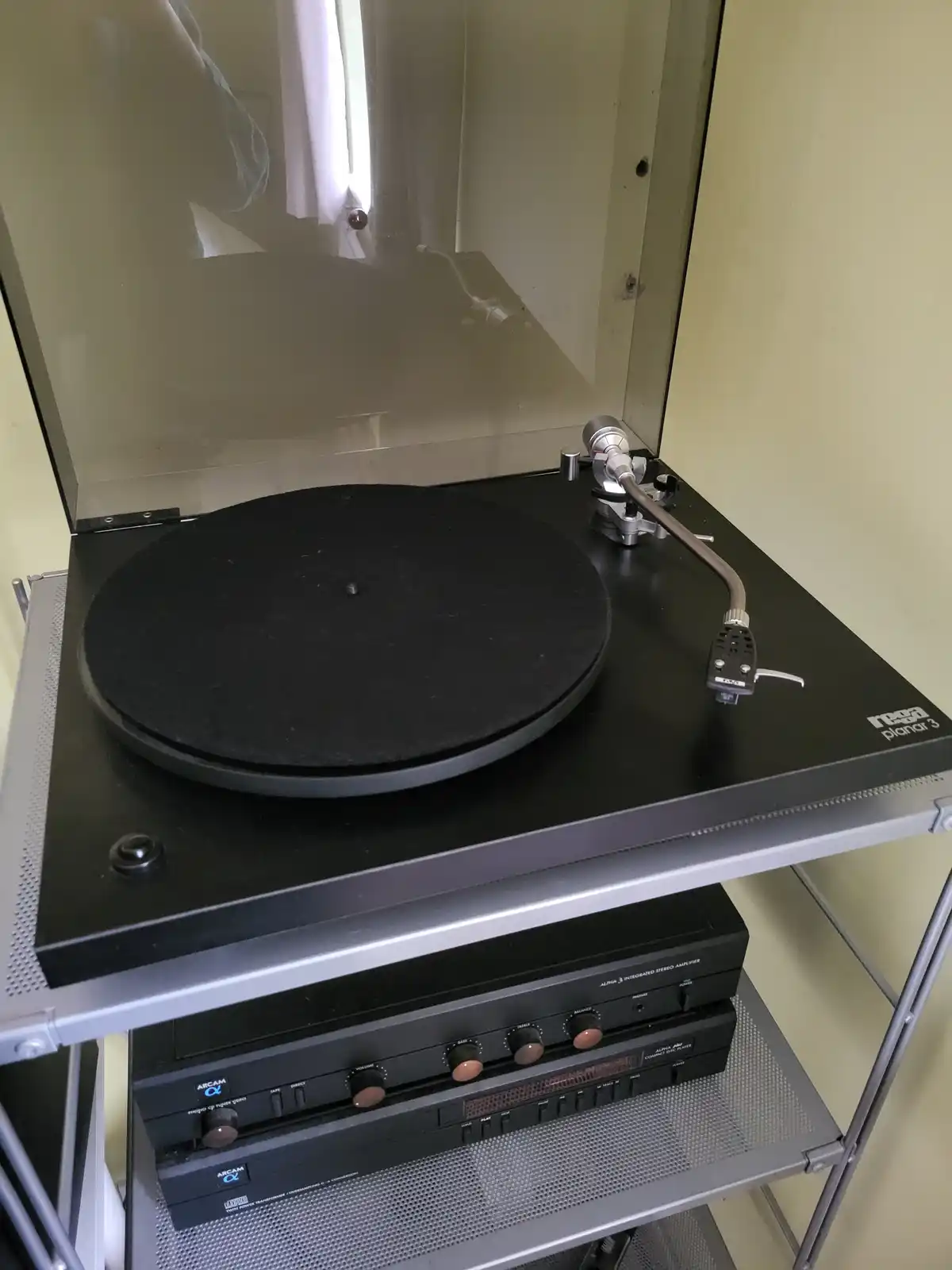 A Rega Planar 3 turntable sits on a metal shelving unit with Arcam audio equipment below, its tonearm poised over an empty…