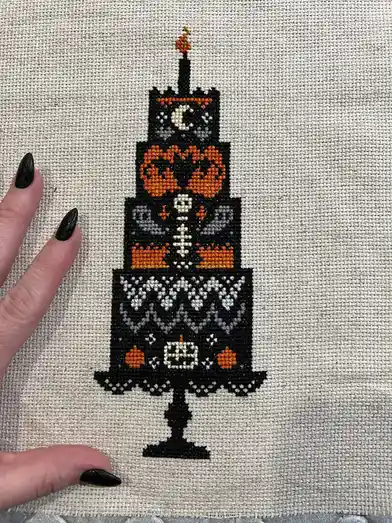 A hand with black nail polish holds a piece of fabric displaying a cross-stitched Halloween-themed cake. The cake is…