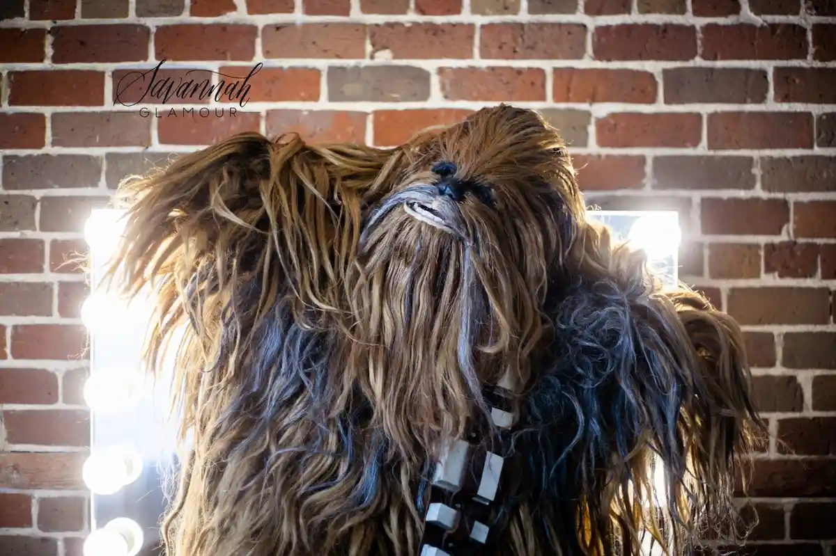 A person in a Chewbacca costume stands before a mirror with bright lights, looking surprised or shocked. The background is a…