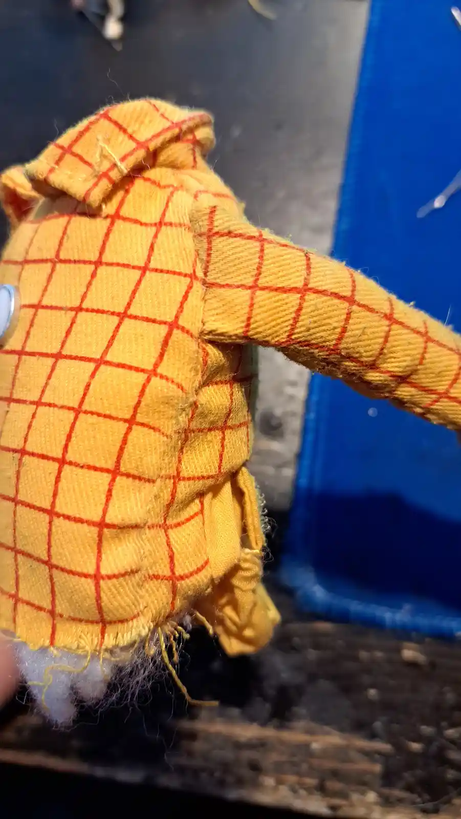 A close-up photo shows the back of a handmade plush toy wearing a yellow and red checkered jacket with frayed edges and…