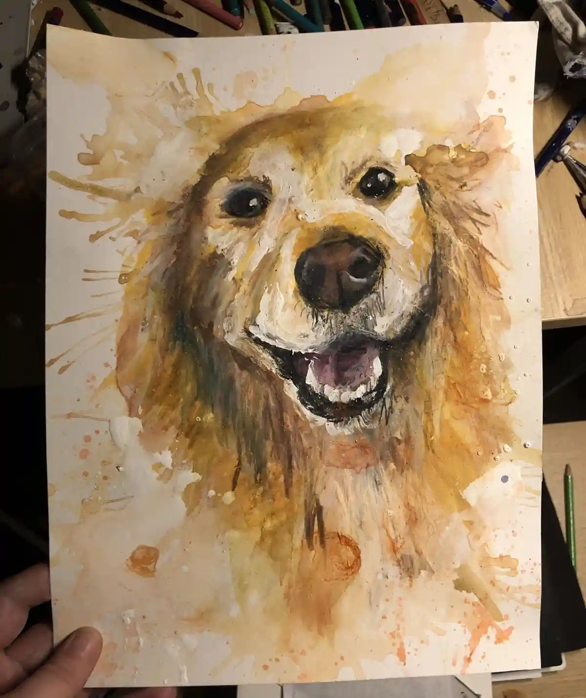 A hand holds up a watercolor painting of a golden retriever with a joyful, open-mouthed expression, surrounded by paint…