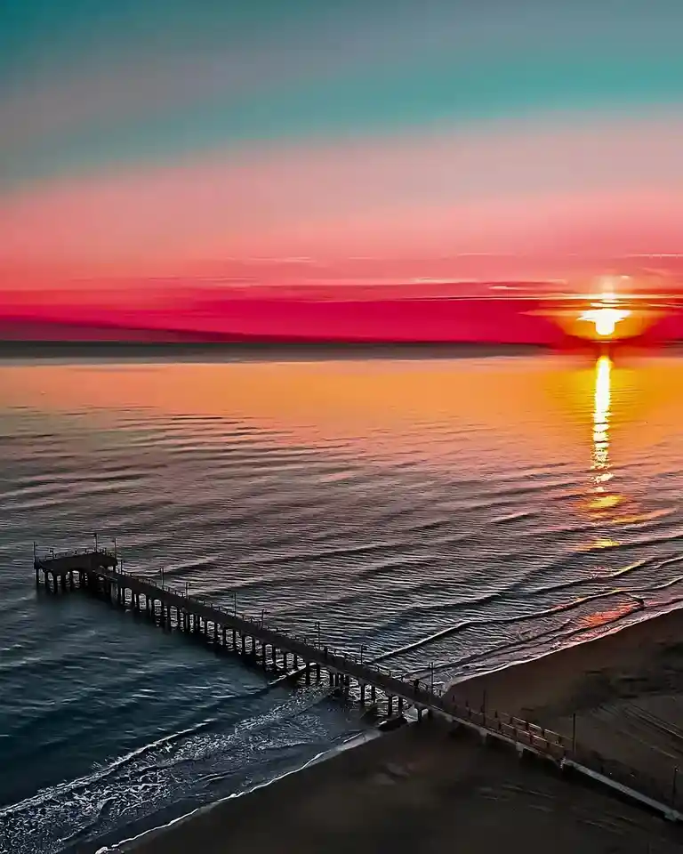 A vibrant sunset over the ocean with a long pier extending into the water, the sky painted in gradients of teal, pink, and…