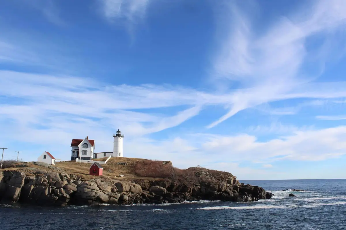 A scenic photo of a white lighthouse and keeper's house on a rocky coastal island under a blue sky with wispy clouds. The…