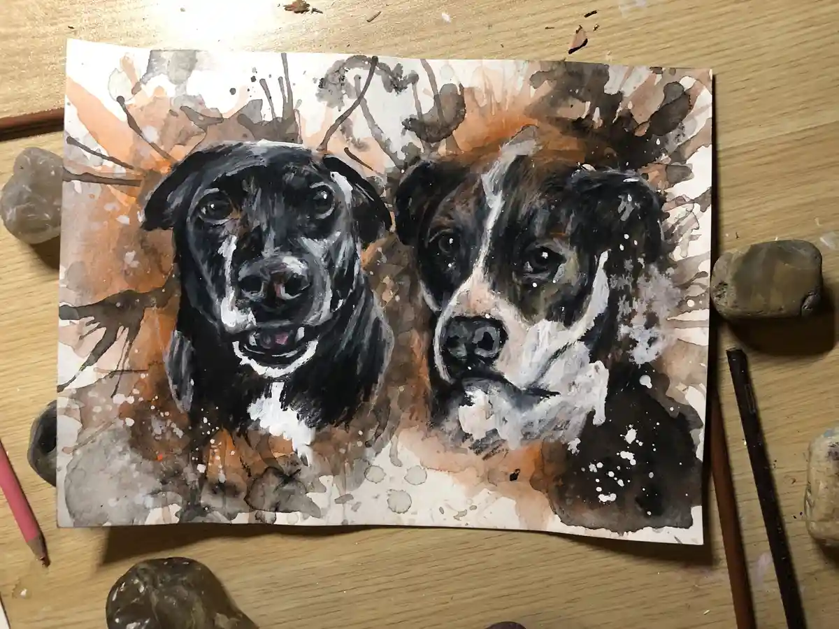 A watercolor painting of two dogs' heads side by side on a piece of paper, resting on a wooden desk surrounded by art…