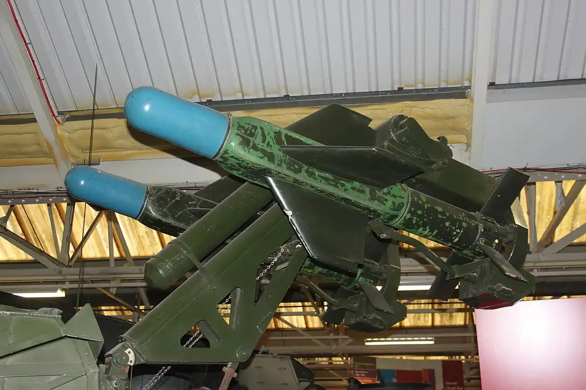 A green military missile with a blue nose cone is mounted on display in a museum, angled upward. The overlaid text reads…