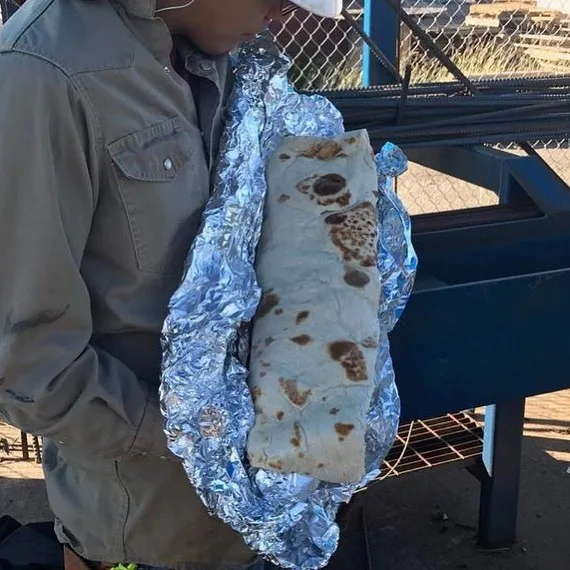 A person wearing a beige jacket and white cap holds a large burrito wrapped in aluminum foil. The burrito is visibly charred…