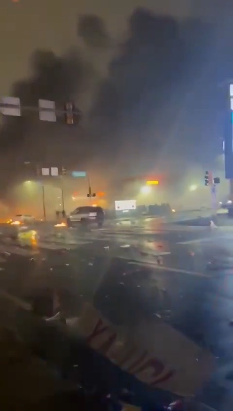 A blurry nighttime street scene viewed from a car, showing thick black smoke billowing into the sky, orange flames visible…