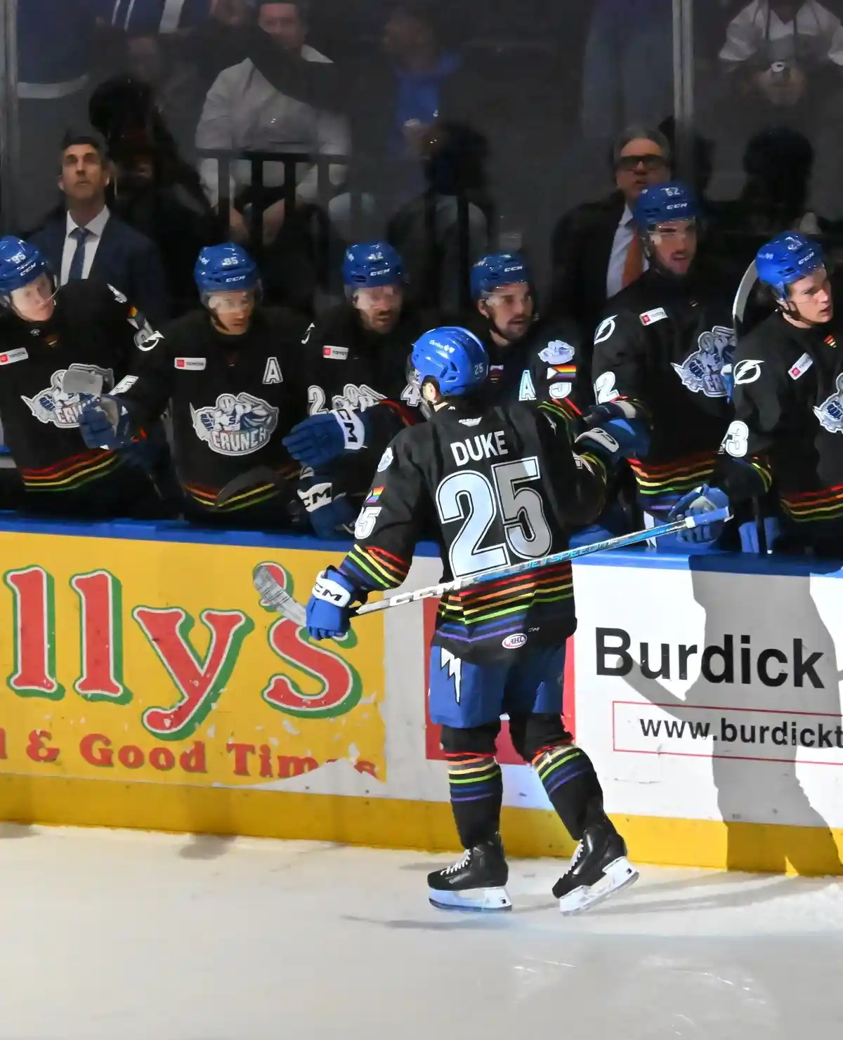 A hockey player wearing a black jersey with 'DUKE 25' on the back and rainbow stripes walks away from the bench while…