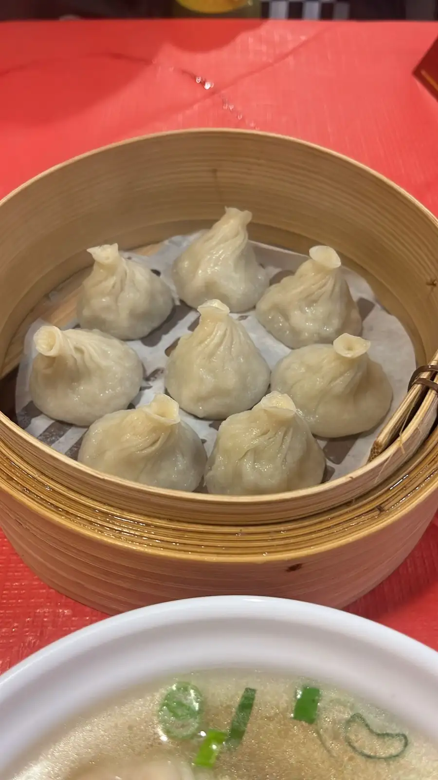 A close-up photo shows a bamboo steamer basket filled with eight plump, steamed dumplings resting on parchment paper, placed…