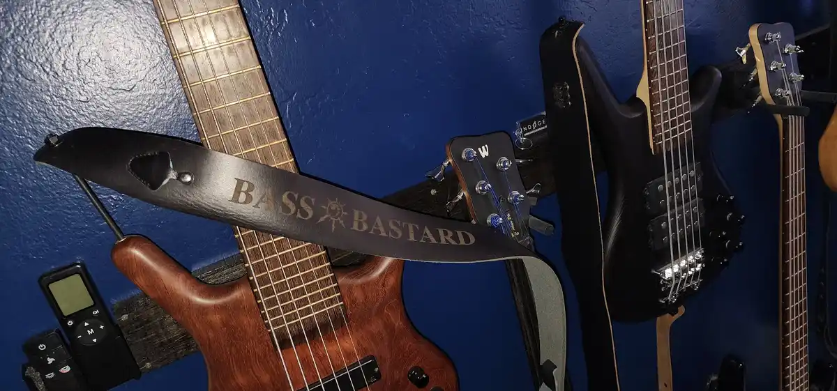 A close-up photo shows a wooden bass guitar with a black leather strap that reads 'BASS BASTARD' in gold lettering, hanging…