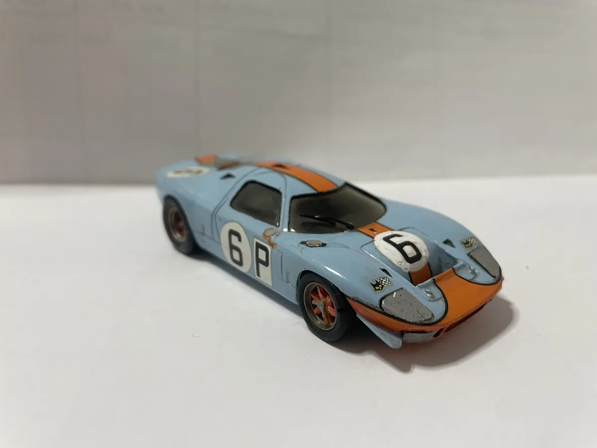 A light blue and orange vintage race car toy, resembling a Ford GT40, is shown on a white surface with visible wear and…