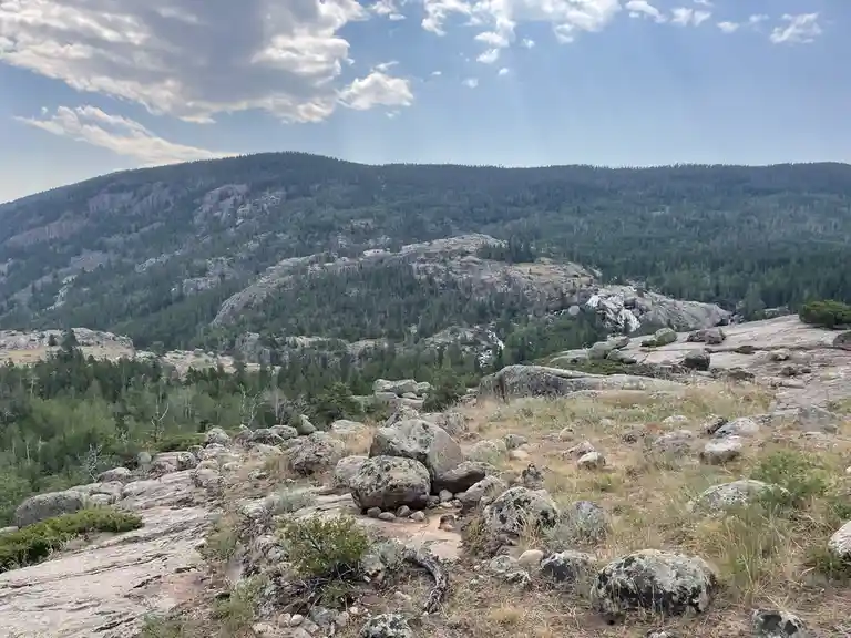 A scenic mountain landscape with rocky terrain, scattered boulders, and a distant waterfall under a partly cloudy sky. The…