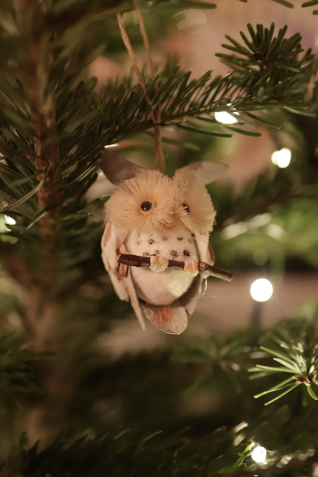 A close-up photo shows a handmade, fluffy white owl ornament hanging from a green pine tree branch, surrounded by…