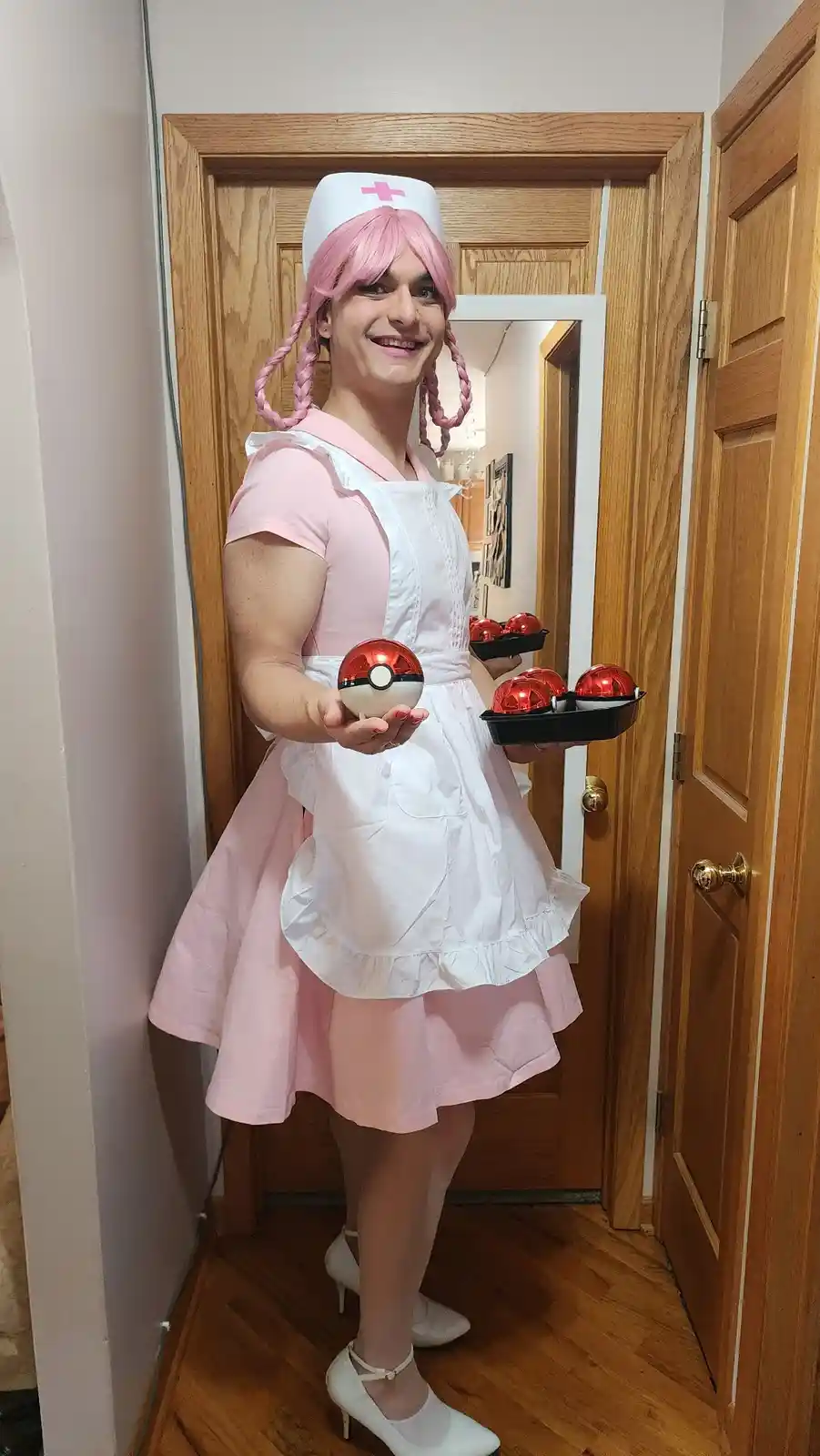 A person in a pink nurse costume with a white apron and pink wig holds a single Poké Ball in one hand and a tray of multiple…