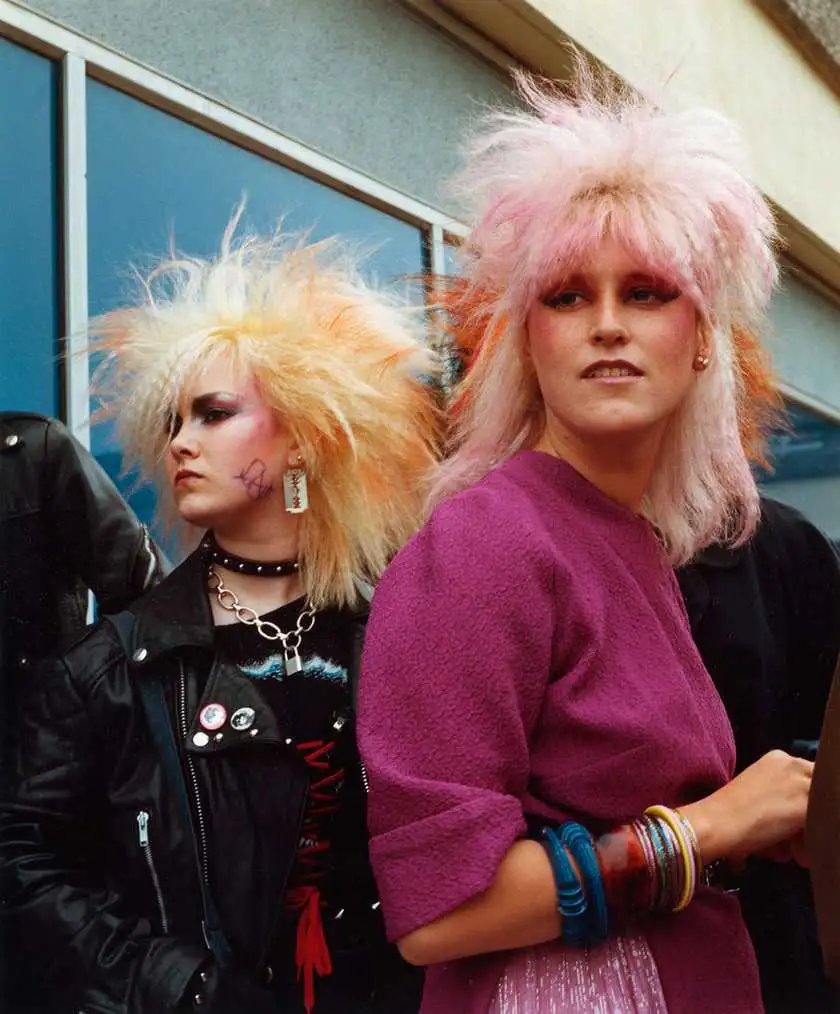 Two women with extreme 1980s punk hairstyles—one with spiky blonde/orange hair and another with fluffy pink hair—pose…