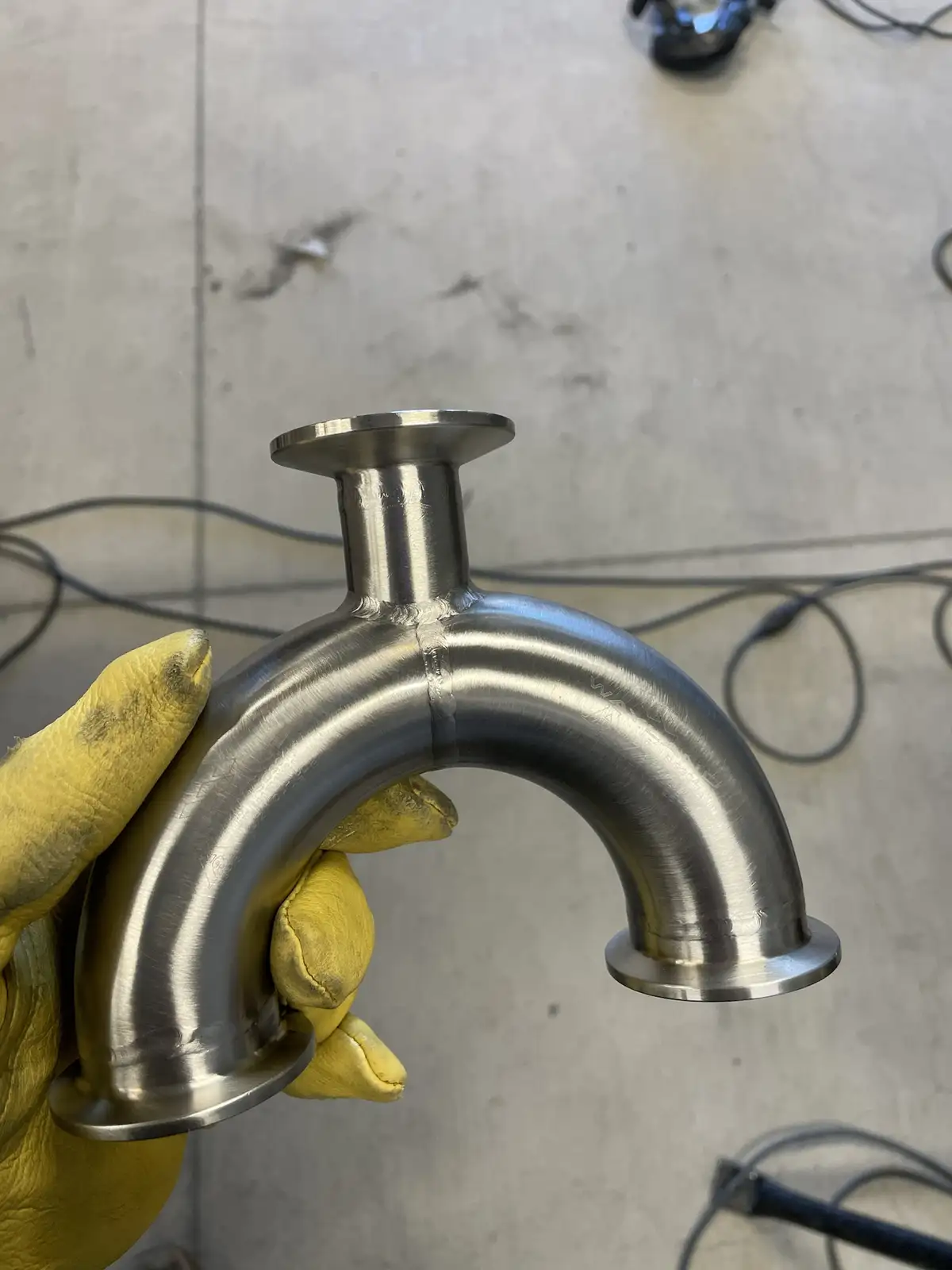 A hand wearing a yellow work glove holds up a shiny, custom-made stainless steel pipe elbow with a visible weld seam. The…