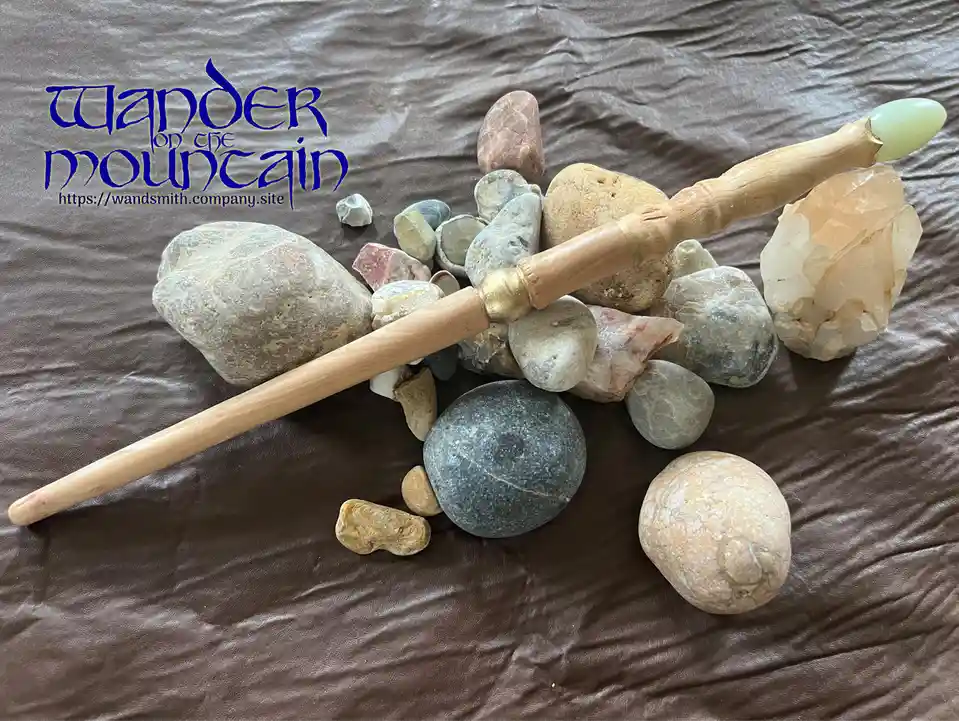 A wooden wand with a green stone tip rests diagonally among a collection of various smooth and rough stones on a brown…