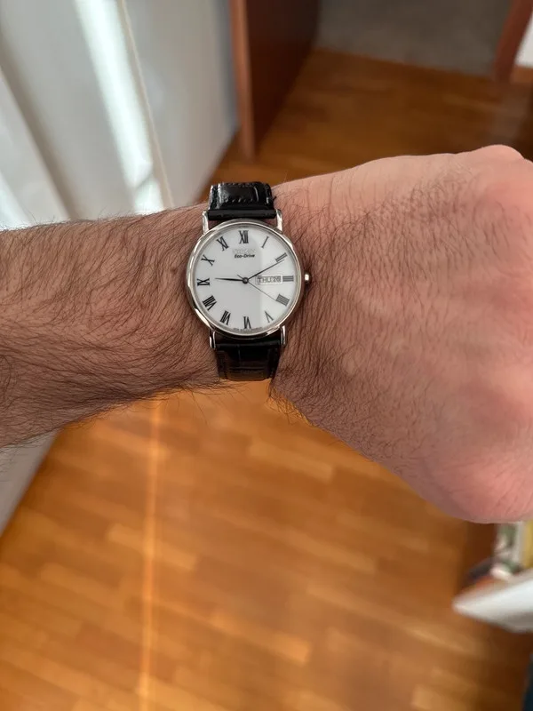 A close-up photo shows a person's hairy left wrist wearing a silver-toned analog watch with a black leather strap. The watch…