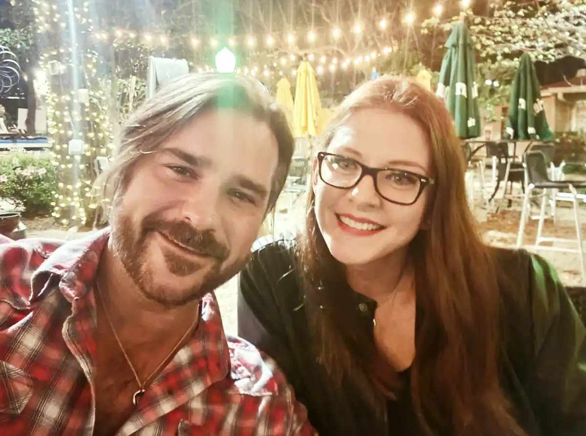 A selfie of a smiling man with a beard and plaid shirt and a smiling woman with glasses, taken at night outdoors under…