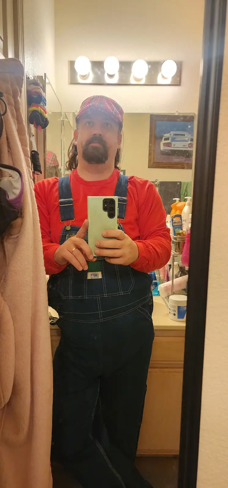 A man with a beard and long hair wearing a red shirt and blue denim overalls, along with a red plaid cap, takes a mirror…
