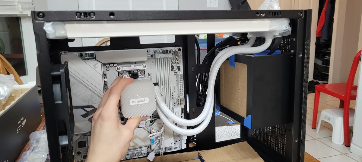 A hand holds a white 'be quiet!' branded CPU water block inside an open black computer case, with white tubing connected to…