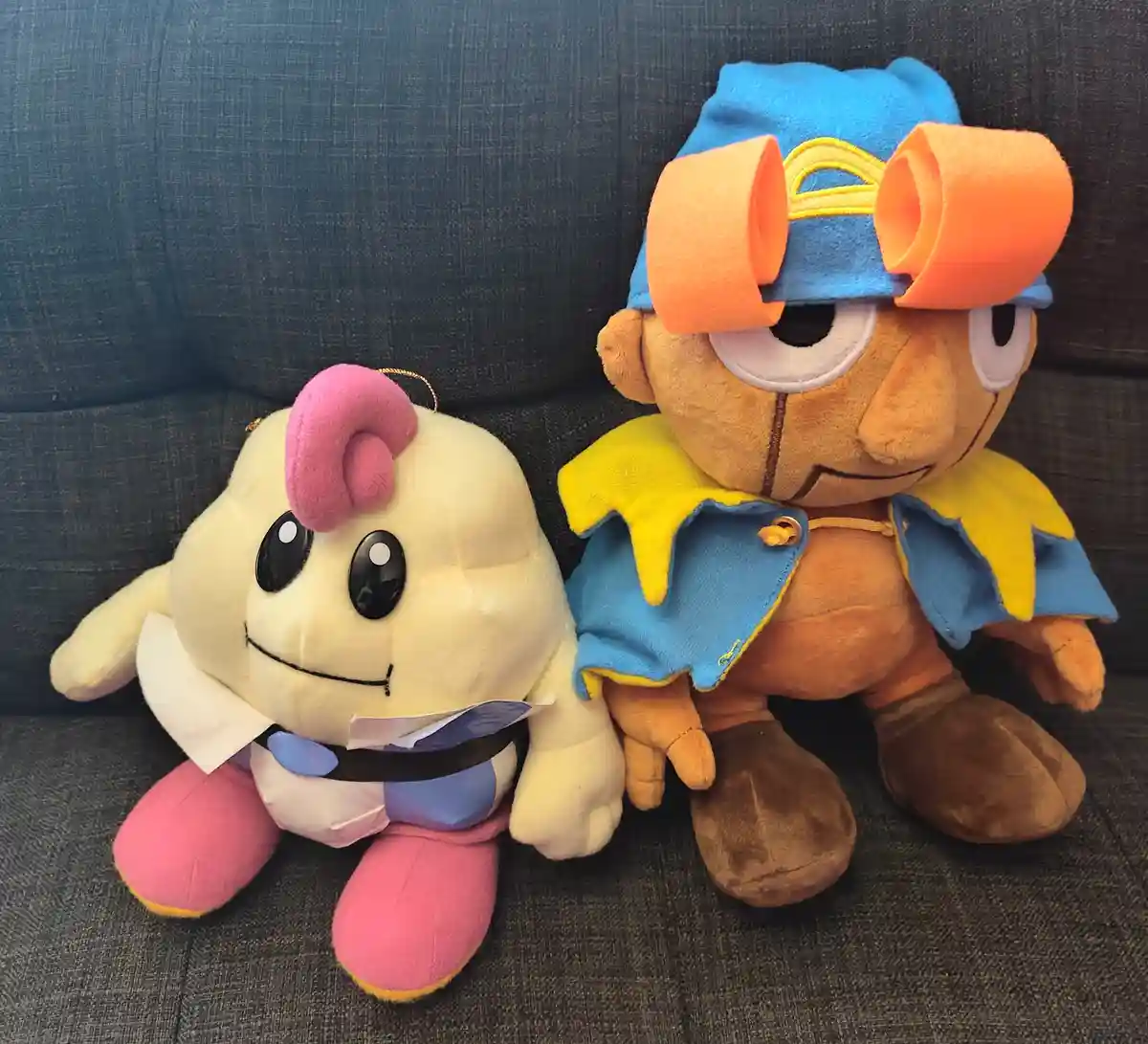 Two plush toys sit side by side on a dark gray couch. The smaller one on the left is cream-colored with a pink mohawk, white…