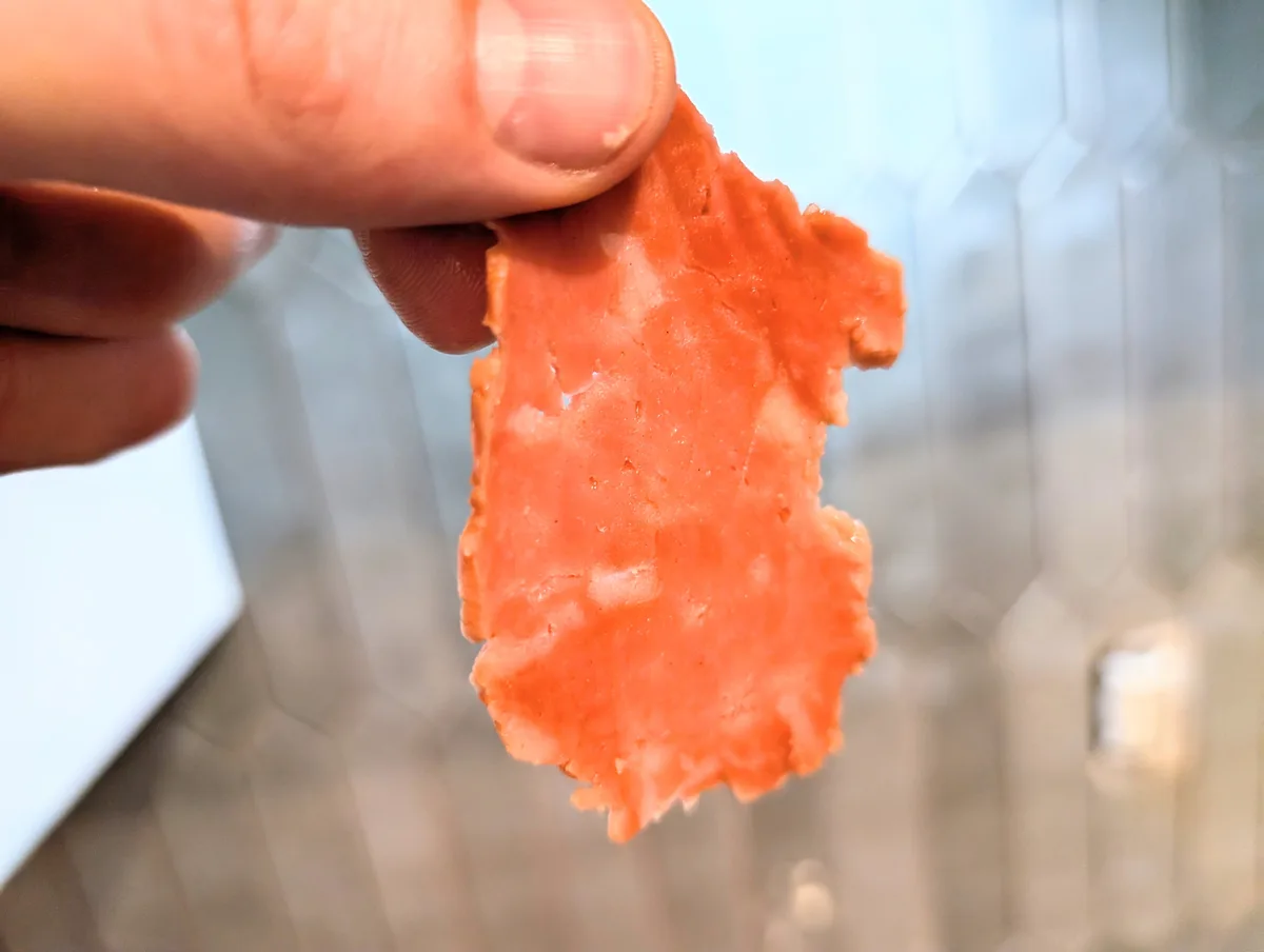 A close-up photo shows a hand holding a thin, orange-red slice of processed meat, possibly a deli slice or meatloaf, with a…