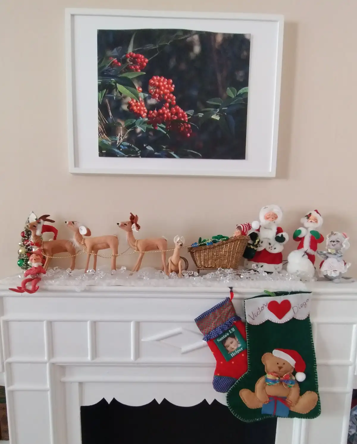A white fireplace mantel is decorated with Christmas figurines including reindeer pulling a sleigh, Santa Claus figures, and…